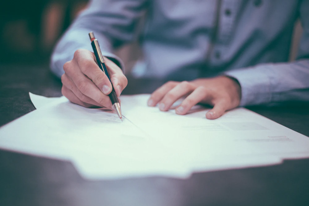 A person signing a document at a desk, pen in hand.